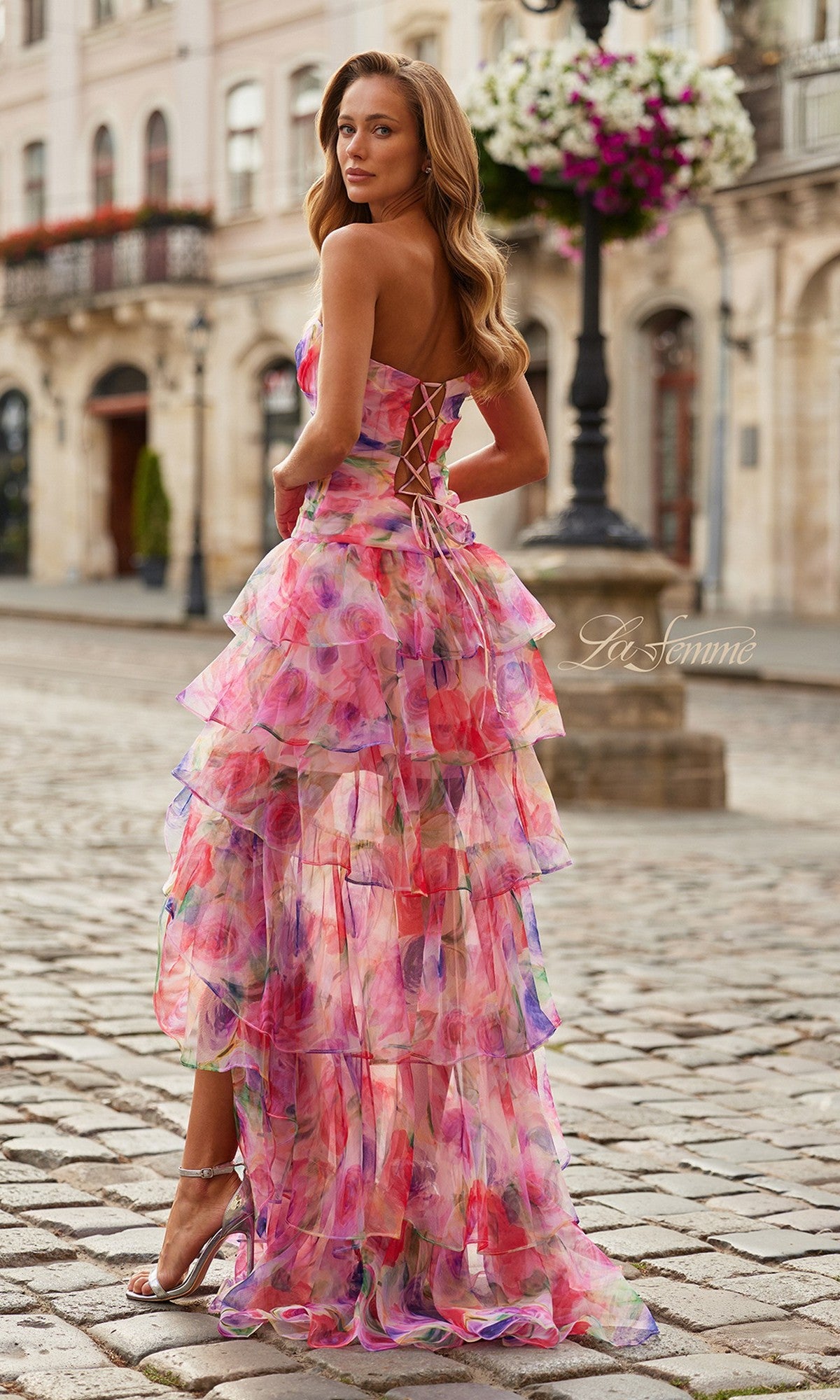 A woman in the Long Prom Dress: La Femme 33555, featuring a colorful tiered floral design and strapless sweetheart neckline, stands on a cobblestone street, glancing over her shoulder.